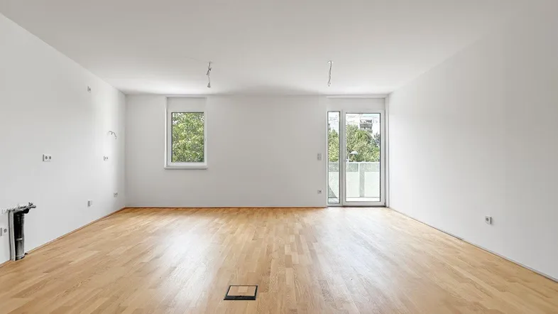 Heller, leerer Wohnraum mit Holzboden, weißen Wänden und Zugang zu einem Balkon.