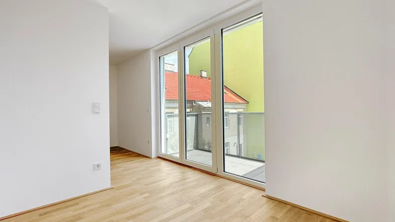 Helles Zimmer mit großen Fenstern, Parkettboden und Zugang zu einem Außenbereich.