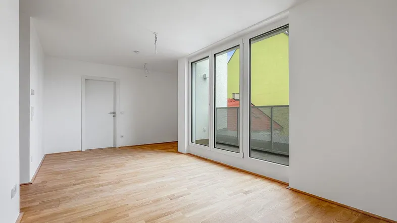 Helles, leeres Wohnzimmer mit Parkettboden und großen Fenstern, die Zugang zu einem Balkon bieten.