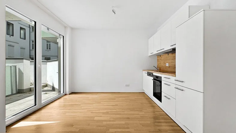 Helle, leere Küche mit modernen weißen Schränken, Holzarbeitsplatte und Zugang zum Balkon.