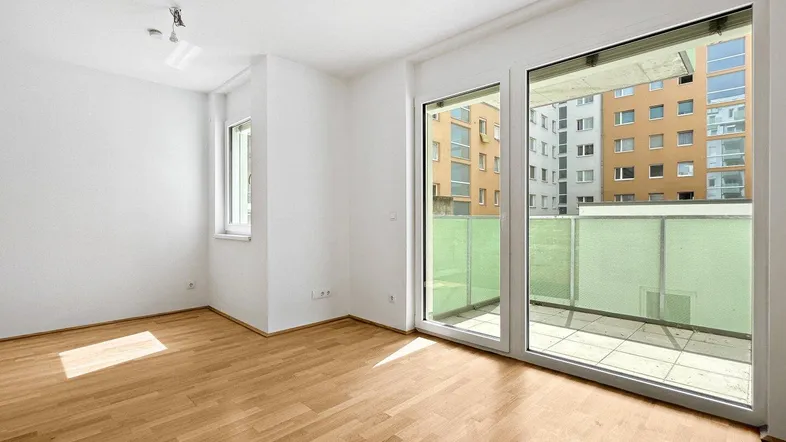 Helles Wohnzimmer mit Holzboden und Zugang zu einem Balkon mit Blick auf umliegende Gebäude.