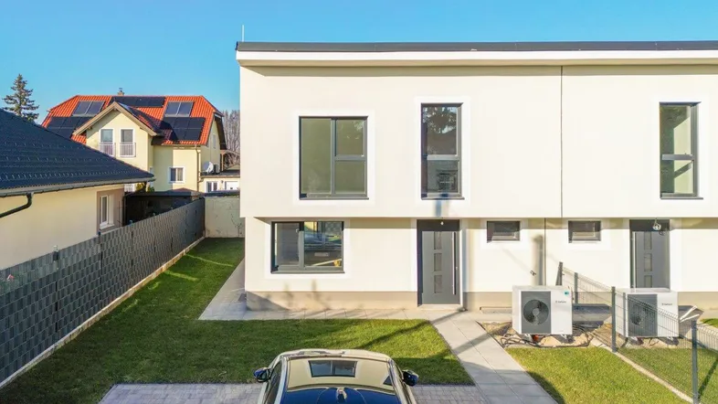 Moderne Hausfassade mit klaren Linien, gepflasterter Einfahrt, Vorgarten und Wärmepumpen an einem sonnigen Tag.