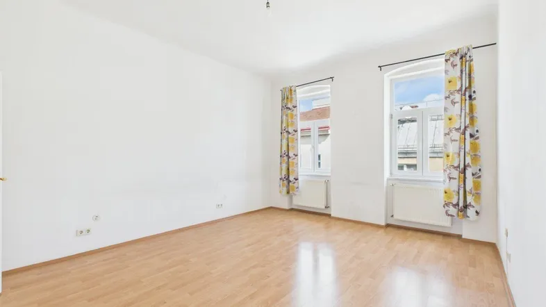 Helles, leeres Zimmer mit Laminatboden und zwei Fenstern, bereit zur individuellen Gestaltung.