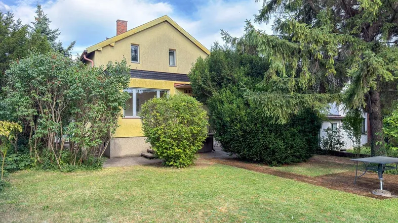 Gelbes Einfamilienhaus mit gepflegtem Vorgarten, Rasenfläche und hohen Bäumen unter blauem Himmel.