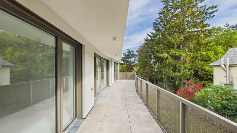 Lange, moderne Terrasse mit Fliesenboden und Metallgeländer, umgeben von üppigem Grün und Bäumen.