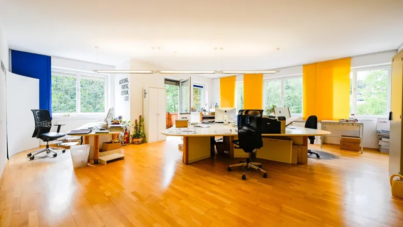 Helles Großraumbüro mit glänzendem Holzboden, mehreren Arbeitsplätzen und großen Fenstern, die viel Tageslicht hereinlassen.