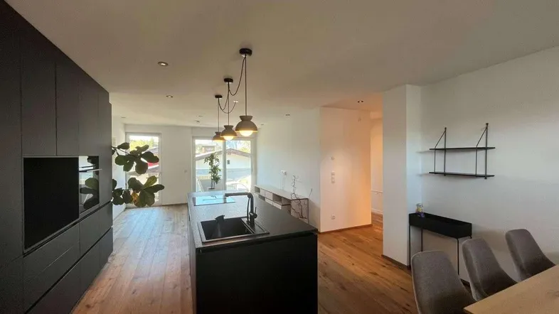 Moderner offener Wohn-Essbereich mit dunkler Küche, Holzboden und Zugang zum Balkon.