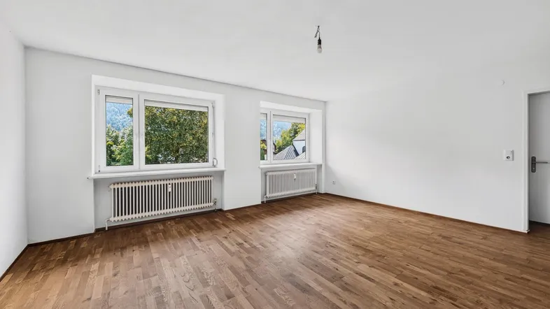 Helles Zimmer mit Holzboden, weißen Wänden und Blick ins Grüne sowie auf die Berge.
