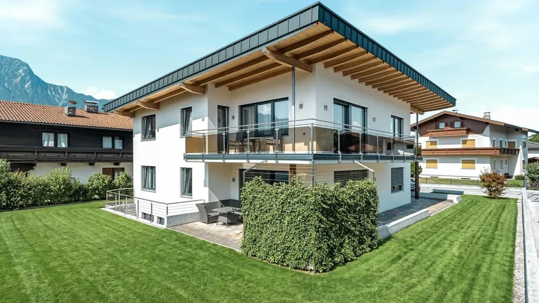 Modernes Einfamilienhaus mit weißer Fassade, großem Garten und Bergblick unter blauem Himmel.