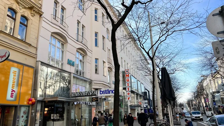 Lebhafte Stadtstraße mit Geschäften, Passanten und historischen Gebäuden unter blauem Himmel.