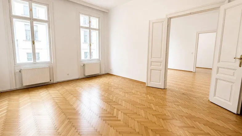 Helles Zimmer mit Fischgrätparkett, hohen Fenstern und Zugang zu weiteren Räumen.
