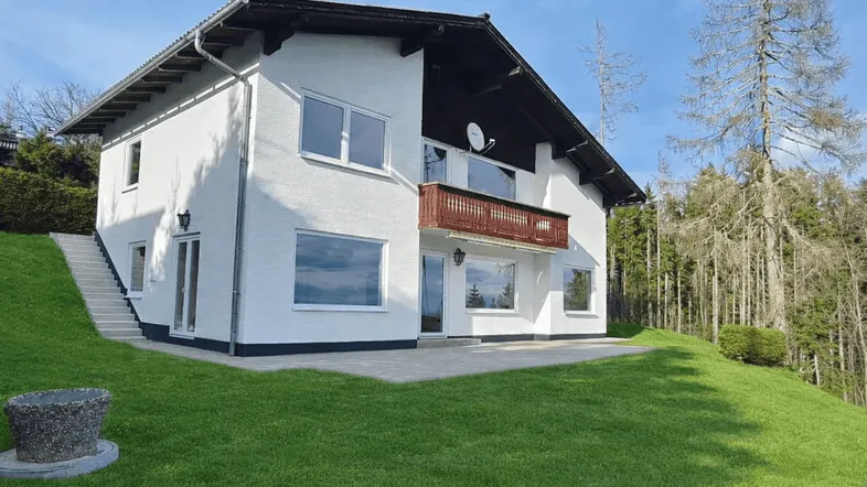 Weißes Einfamilienhaus mit Satteldach, Balkon und großer Rasenfläche in bergiger Umgebung.