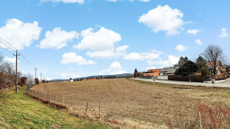 Weites Feld mit ländlicher Umgebung und Häusern im Hintergrund unter blauem Himmel.