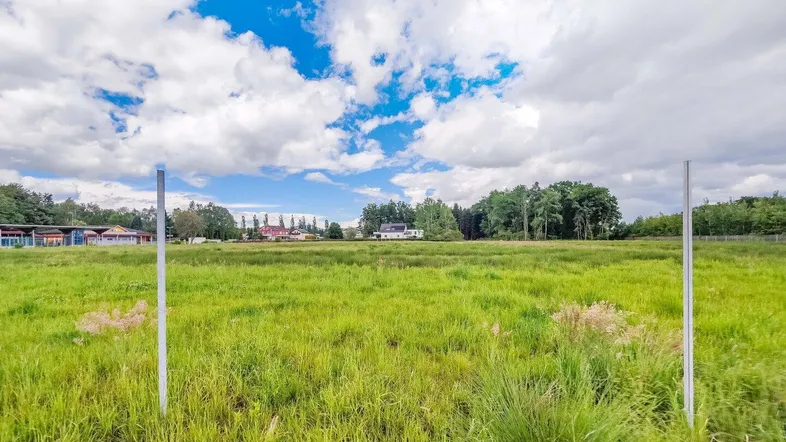 Weitläufiges grünes Feld unter bewölktem Himmel mit Bäumen und Gebäuden in der Ferne.