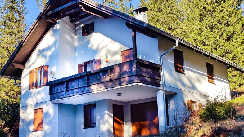 Traditionelles Einfamilienhaus mit Satteldach und Holzbalkon, umgeben von Nadelbäumen in sonniger Lage.