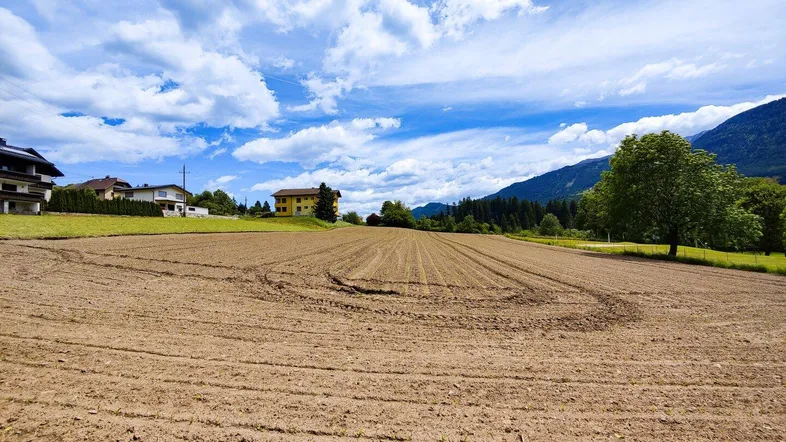 Weitläufiges Ackerland mit Häusern im Hintergrund und majestätischen Bergen unter blauem Himmel.