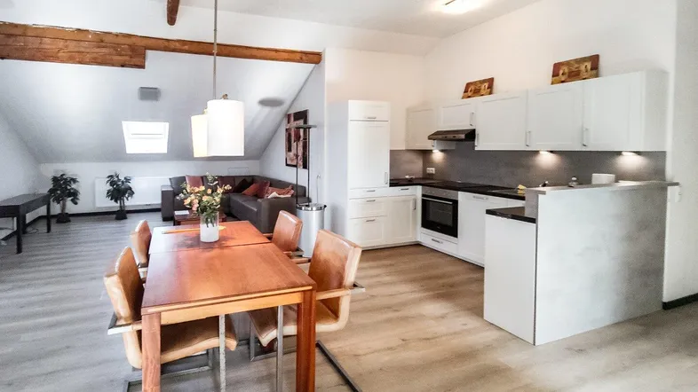 Helles Dachgeschoss-Wohnzimmer mit Essbereich, offener Küche und sichtbaren Holzbalken.