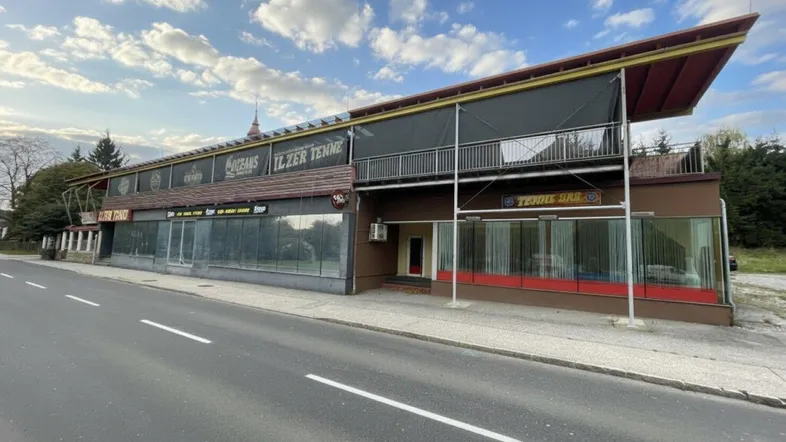 Gebäudeansicht von der Straße mit großen Schaufenstern und einem Vordach unter bewölktem Himmel.