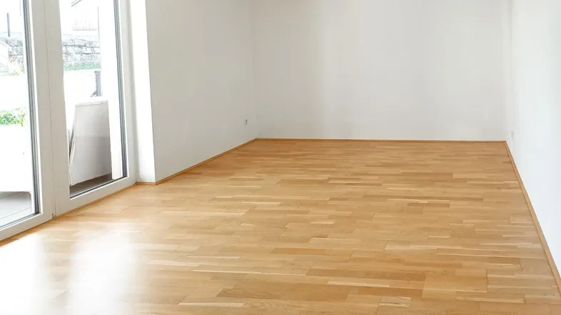 Helles, leeres Zimmer mit Parkettboden und Zugang zu einem Balkon oder einer Terrasse.