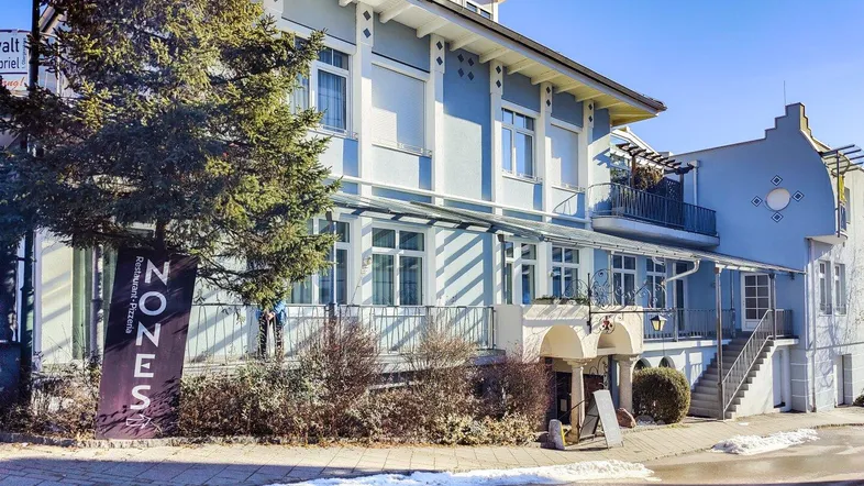 Ein charmantes, hellblaues Gebäude mit mehreren Etagen, großen Fenstern und einem kleinen Balkon, umgeben von Bäumen und etwas Schnee.
