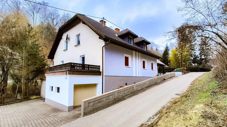 Ansicht eines Einfamilienhauses mit Balkon und Auffahrt, umgeben von grüner Natur und Bäumen.