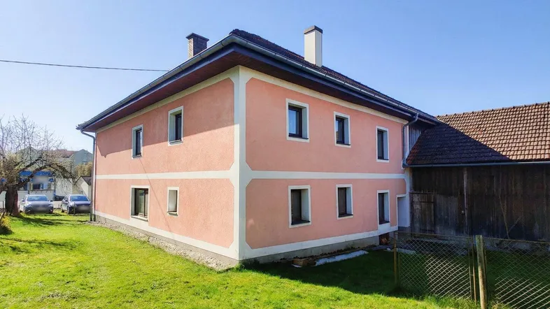 Rosa verputztes Wohnhaus mit Satteldach und grünem Rasen unter blauem Himmel.