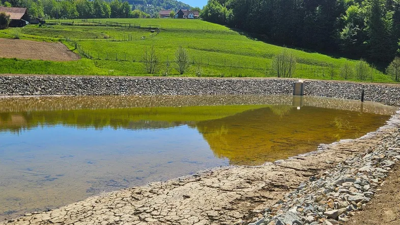 Ein künstlich angelegter Teich oder Wasserspeicher in einer ländlichen Umgebung mit grünen Hügeln und Bäumen im Hintergrund.
