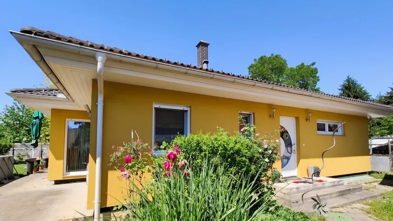 Gelbe Hausfassade mit braunem Ziegeldach und gepflegtem Vorgarten unter blauem Himmel.