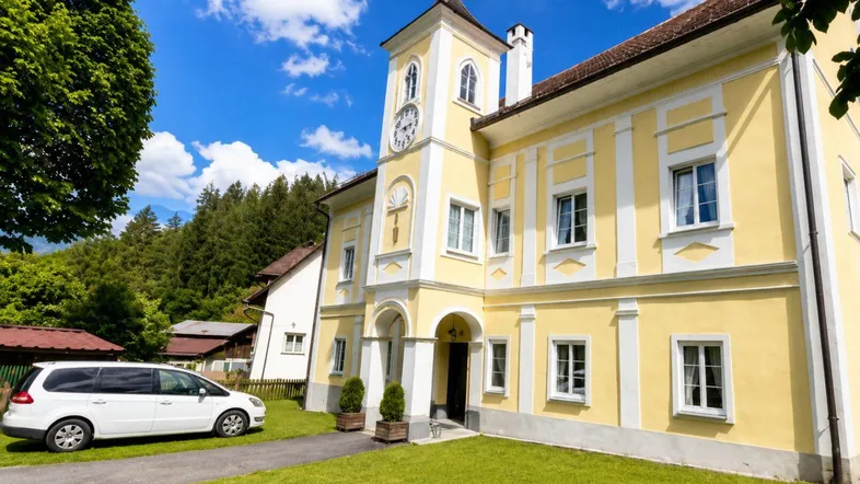 Historisches Gebäude mit gelber Fassade, Uhrturm und gepflegtem Vorgarten unter blauem Himmel.