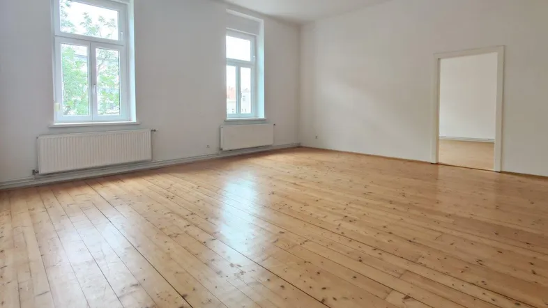 Helles, leeres Zimmer mit Holzdielenboden und zwei großen Fenstern, die viel Tageslicht hereinlassen.