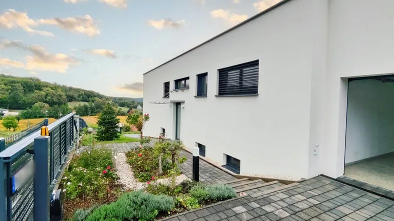 Moderne weiße Fassade eines Hauses mit gepflegtem Vorgarten und Blick auf die umliegende Landschaft.