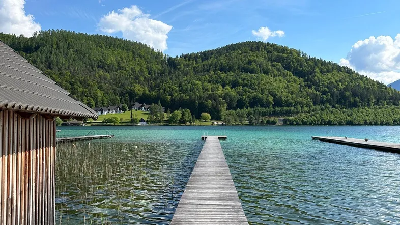 Langer Holzsteg führt in einen klaren See mit bewaldeten Hügeln im Hintergrund und einem Bootshaus.