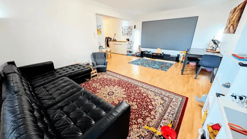 Geräumiges Wohnzimmer mit schwarzem Ledersofa, Esstisch und Projektionsfläche für Unterhaltung.
