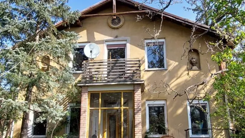 Altes Haus mit verputzter Fassade, Balkon und umgebender Vegetation unter blauem Himmel.