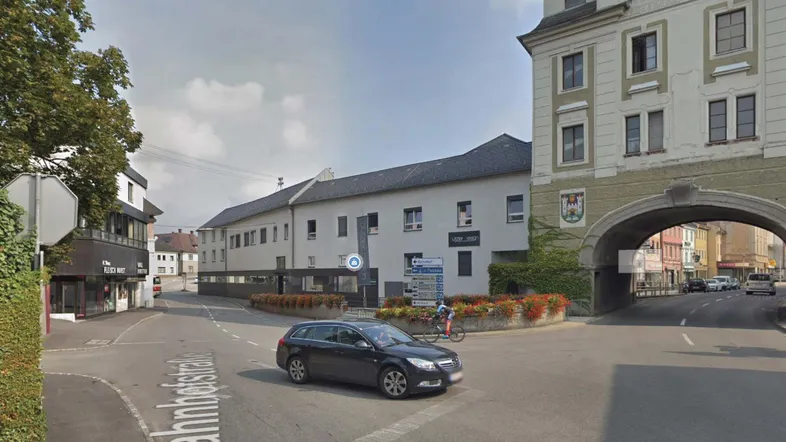 Straßenansicht mit Gebäuden, einem Auto und einem historischen Torbogen in einer städtischen Umgebung.