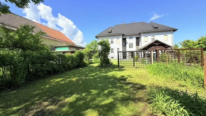 Grüner Gartenbereich mit Rasenfläche und Hecken, umgeben von einem Zaun und Gebäuden.