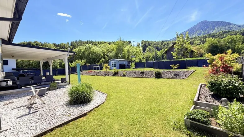 Gepflegter Garten mit großer Rasenfläche, überdachter Terrasse und Bergblick unter blauem Himmel.