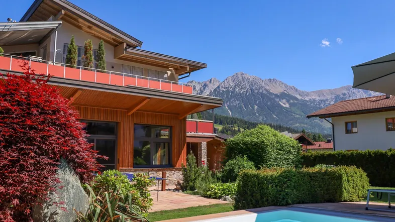 Modernes Haus mit Pool und Garten, umgeben von einer beeindruckenden Berglandschaft unter blauem Himmel.
