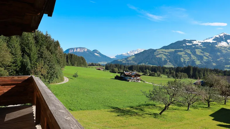 Blick vom Holzbalkon auf eine weite grüne Landschaft mit majestätischen Bergen im Hintergrund unter blauem Himmel.