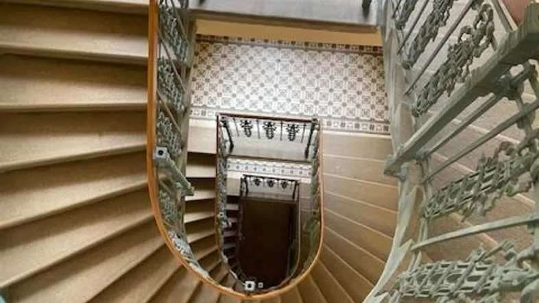 Blick von oben auf ein historisches Treppenhaus mit kunstvollem Geländer und gemusterten Fliesen.