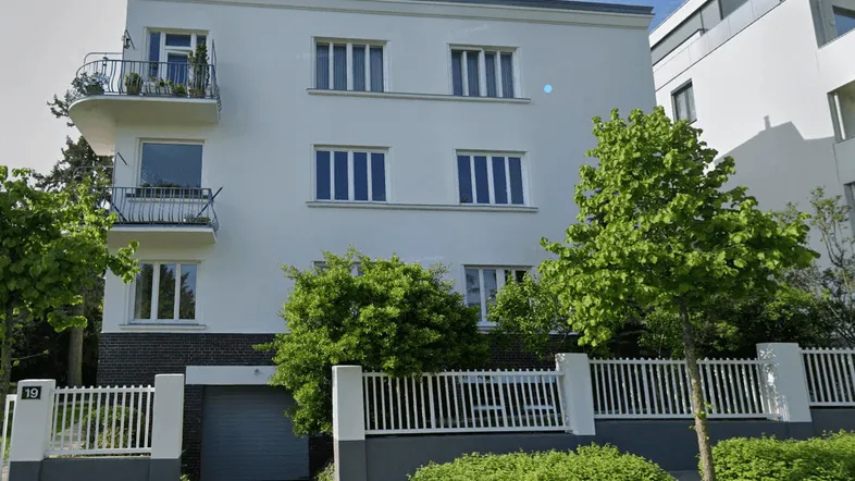 Weißes Mehrfamilienhaus mit modernen Balkonen und einer Garage, umgeben von Grünflächen und einem Zaun.