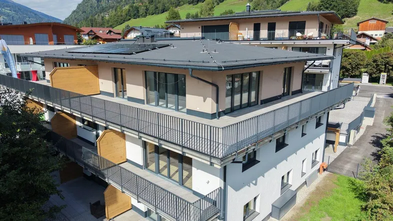 Modernes Mehrfamilienhaus mit mehreren Balkonen, Flachdach und Photovoltaikanlage vor alpiner Bergkulisse.