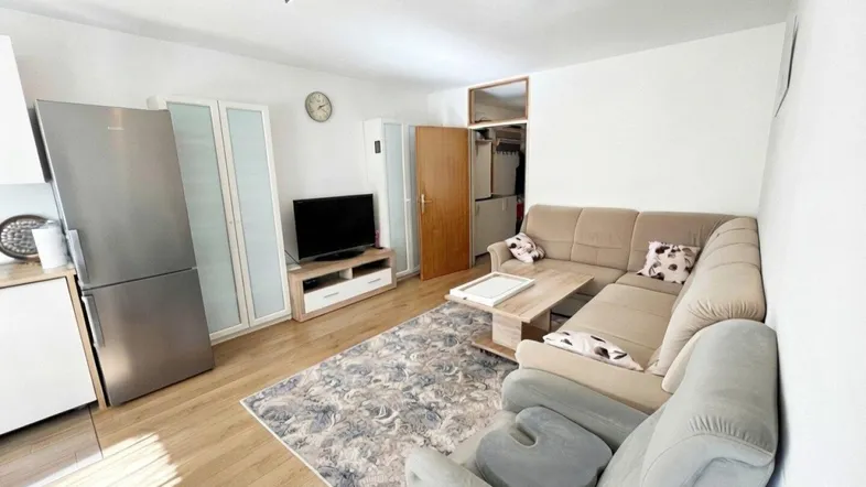 Helles Wohnzimmer mit beigem Ecksofa, Holzboden und integrierter Küchenzeile.