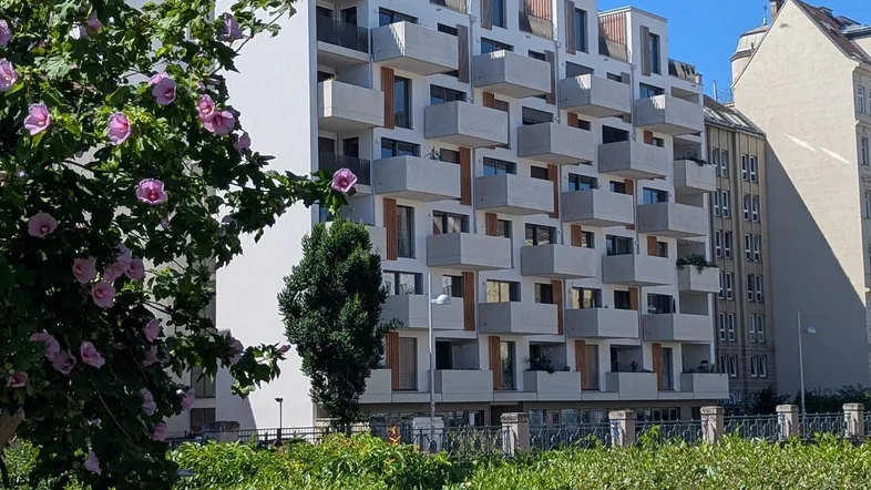 Modernes Apartmentgebäude mit vielen Balkonen, umgeben von grünen Hecken und blühenden Sträuchern.