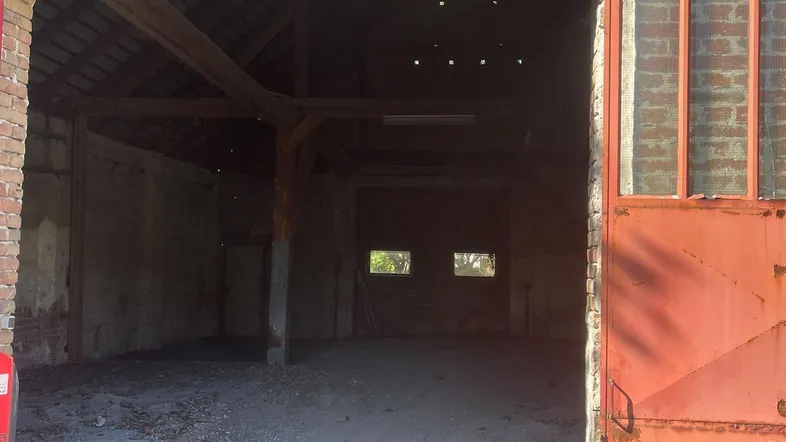Blick in eine große, leere Lagerhalle mit Ziegelwänden, Holzbalken und einem Schotterboden.