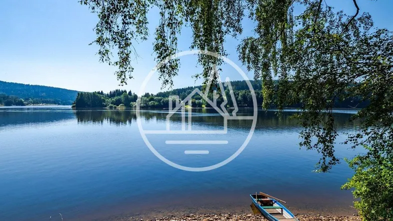 Idyllischer Seeuferblick mit kleinen Booten am Ufer und grüner Vegetation unter blauem Himmel.