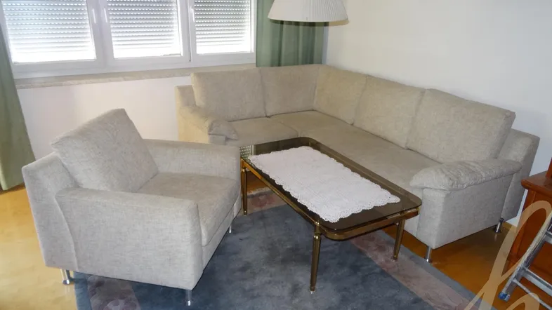 Helles Wohnzimmer mit beigem Ecksofa, Sessel, Couchtisch und blauem Teppich vor dem Fenster.