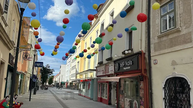 Eine belebte Einkaufsstraße mit bunten Laternen, die über die Gasse gespannt sind, unter blauem Himmel.