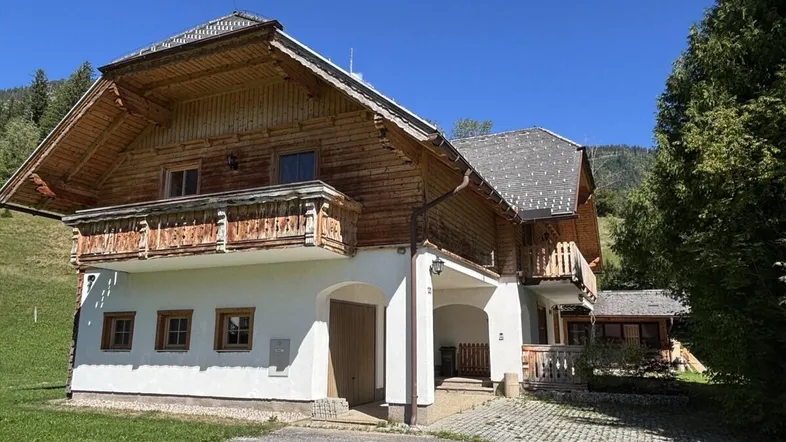 Traditionelles Holzhaus im alpinen Stil mit Balkonen und weißem Sockel in grüner Umgebung.