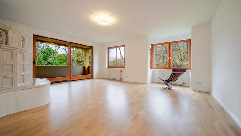 Helles Wohnzimmer mit Parkettboden, Kachelofen und Zugang zu einem großen Balkon mit Waldblick.